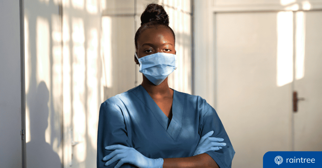 A Healthcare Worker Wearing A Blue Surgical Masks And Gloves, With Their Hair In A High Bun, Looks At The Camera With Their Arms Crossed. Illustrating The Concept Of &Quot;Irrepressible Post-Pandemic Healthcare Trends.&Quot;