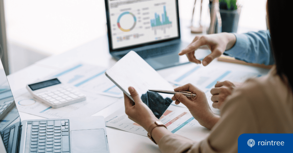 Two Healthcare Administrators Down At A Desk, Which Holds Multiple Devices And Data Spreadsheets. Illustrating The Topic: &Quot;What Is Business Intelligence?&Quot;