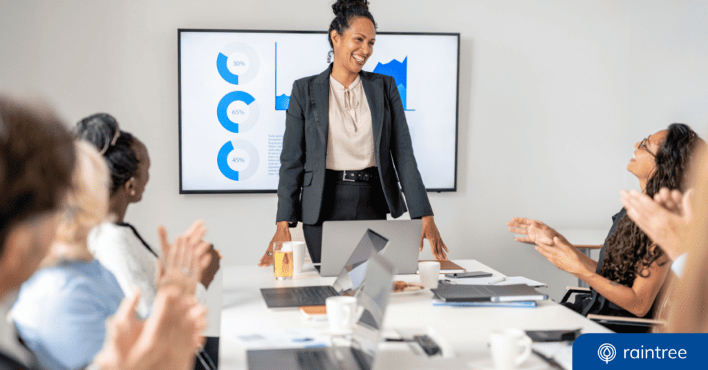 A Physical Therapy Practice Ceo Stands At The Head Of A Desk, Smiling At Her Colleagues. Illustrating The Topic: &Quot;Intro To Business Intelligence For Healthcare Practices.&Quot;