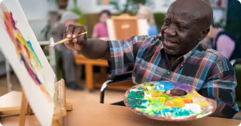 A man in a wheelchair holding a paint brush and a palette. Illustrating the impact of occupational therapy month.