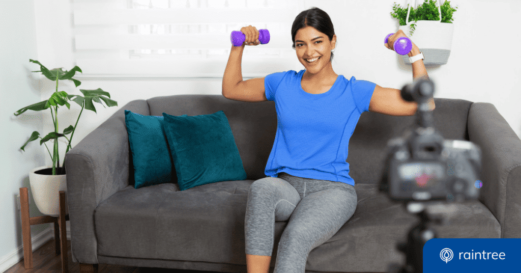A person wearing exercise clothing sits on a gray couch holding two purple dumbbell weights, looking at a camera on a tripod and smiling. Illustrating the topic: "How to Grow Your Small Practice with Social Media."