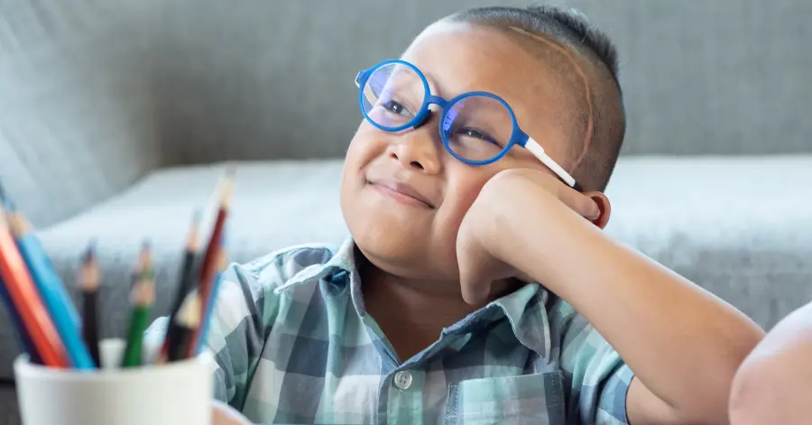 Occupational Therapy and Traumatic Brain Injuries. A header image depicting a child who looks up from a table, where they sit, drawing. They wear pediatric glasses and have a large scar on their temple, indicating traumatic brain injury.