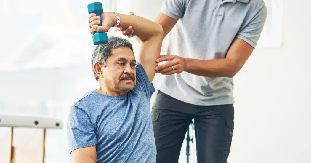 A physical therapist works with a patient, who holds a single dumbbell above his head; illustrating the topic of compliant EMR software.