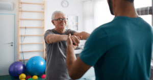 A patient with white hair and glasses smiles at a physical therapist. Illustrating the topic of the importance of sharing patient health information for rehabilitative therapy success.