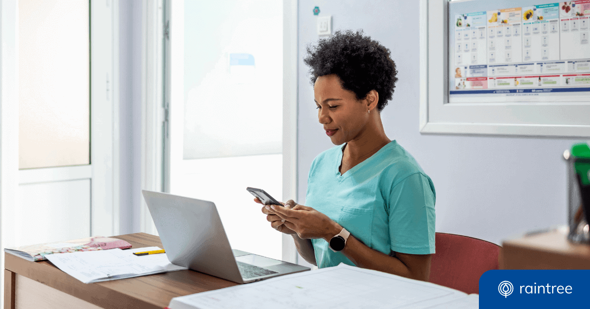 A physical therapist wearing light green scrubs sits at a desk, looking down at a cell phone, with an open laptop and paperwork nearby. Illustrating the topic of "healthcare data security tips."