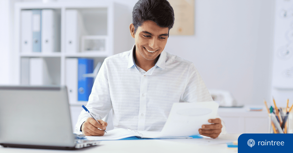 A smiling person wearing a white button up is sitting at a desk and looking down at a paper, while holding a pen. There is a laptop on the desk, in the foreground. Illustrating the topic of password best practices for healthcare organizations.