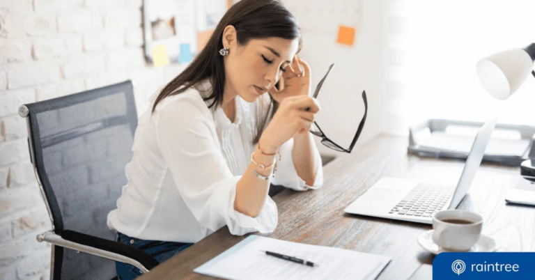 A financial executive sits at a desk with one hand on her forehead and the other holding a pair of glasses, closing her eyes as if frustrated. Illustrating the topic, "Warning Signs Your Therapy Practice Revenue Is at Risk"