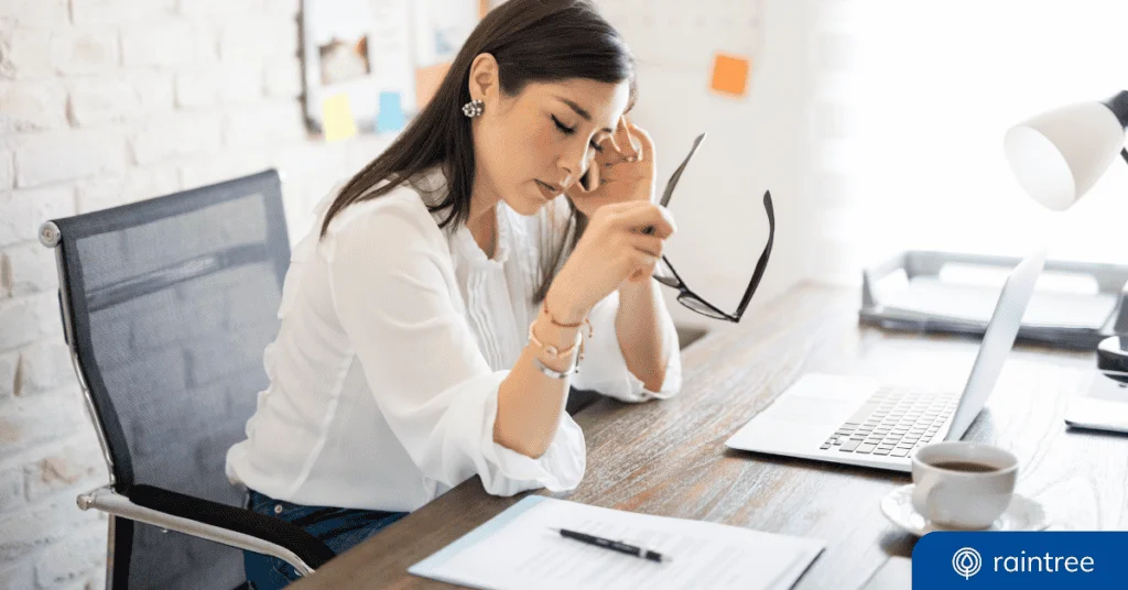 A financial executive sits at a desk with one hand on her forehead and the other holding a pair of glasses, closing her eyes as if frustrated. Illustrating the topic, "Warning Signs Your Therapy Practice Revenue Is at Risk"