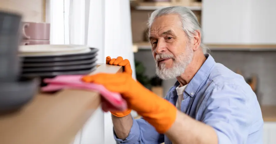 A Senior Man Wearing Orange Gloves Wipes A Shelf, Illustrating The Topic: &Quot;Medicare Billing And Occupational Therapy.&Quot;