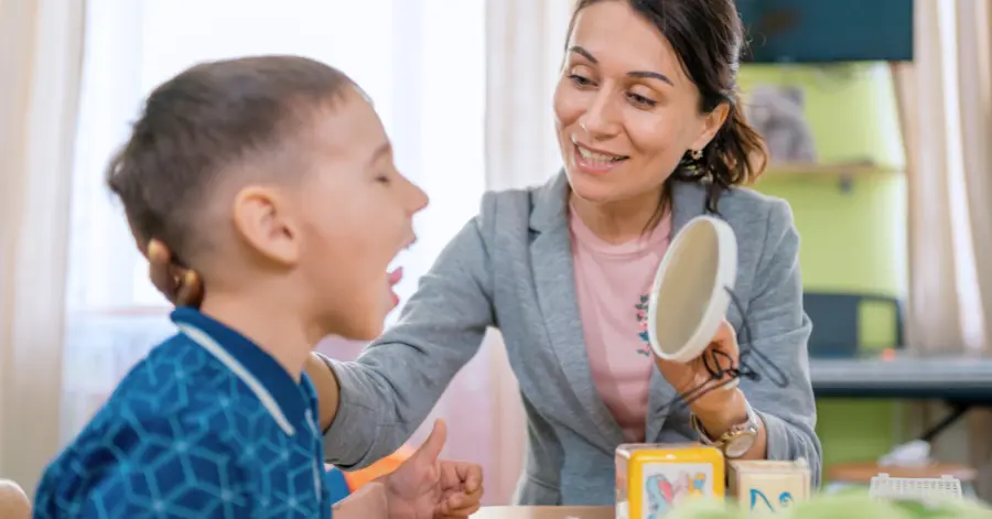 A Speech Therapist Works With A Preschool Aged Boy, Who Is Sticking His Tongue Upward And Looking Into A Mirror. Illustrating The Topic Of Speech Therapy Cpt Codes.