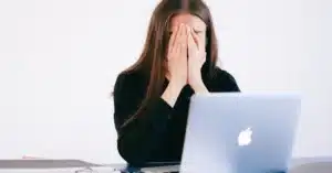 A therapy practice admin with her hand covered by her face, sitting at a computer - illustrating the topic of frustration due to costly EMR downtime.