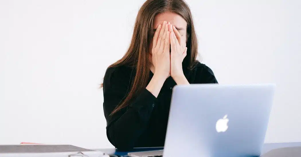 A therapy practice admin with her hand covered by her face, sitting at a computer - illustrating the topic of frustration due to costly EMR downtime.