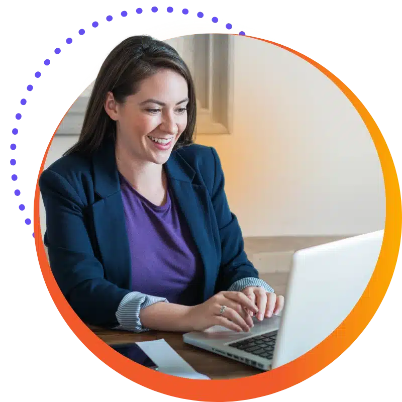Contact Us 1 Contact Us Or Get In Touch Hero: A Professional With Long Brown Hair And A Purple Shirt Sits At A Desk, Smiling Down At Her Laptop.