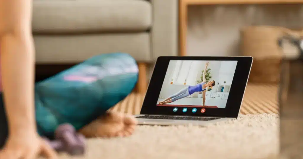 A physical therapy patient sits cross-legged on the floor while watching a home exercise program mobile app video, illustrating the topic of HEP compliance strategies.