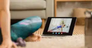 A physical therapy patient sits cross-legged on the floor while watching a home exercise program mobile app video, illustrating the topic of HEP compliance strategies.