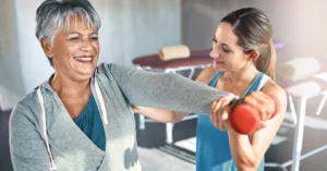 A physical therapist supports the arm of a patient who holds a dumbbell in front of her, smiling; illustrating the topic of ONC certification for rehab therapy and PT software.