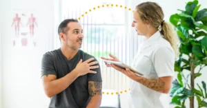A physical therapist holding a tablet makes eye contact with a PT patient who touches his shoulder; behind them is a sound wave, illustrating the benefits of ambient AI in the rehabilitation therapy setting.